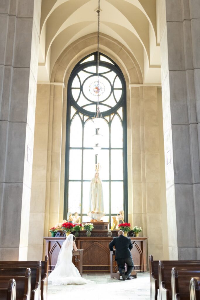 Catholic bride and groom praying at kneelers in front of a statue of Our Lady of Fatima.