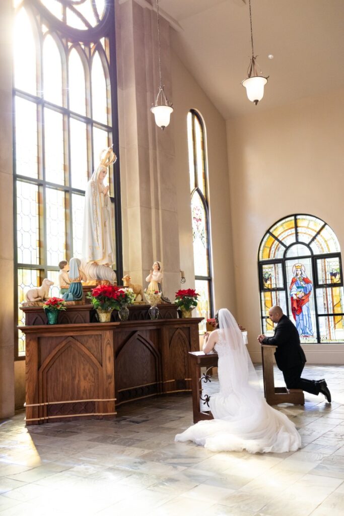 Catholic bride and groom praying at kneelers in front of a statue of Our Lady of Fatima.